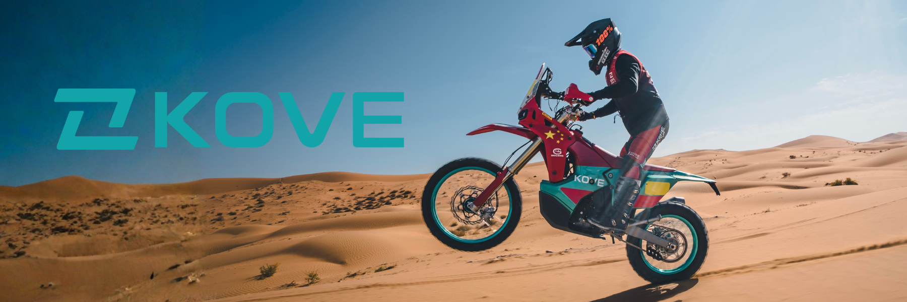 A rider performs a wheelie on a colorful off-road motorcycle in a vast desert landscape with sand dunes.