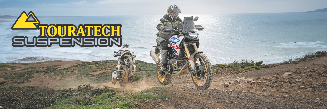 A motorcycle riding on a dirt path near the coast, with two riders in the background. The image features the Touratech Suspension logo prominently on the top left.