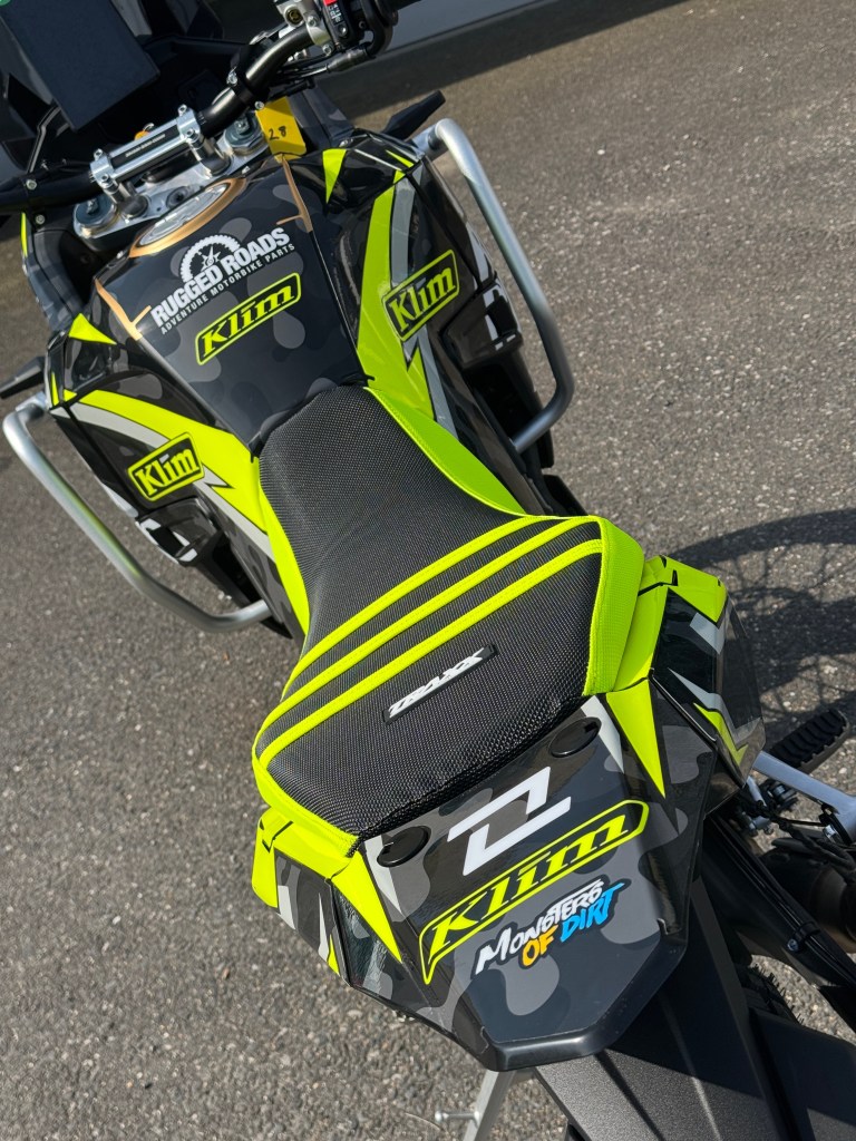 Close-up view of a black and yellow off-road motorcycle, featuring a textured seat and branding from Klim and Rugged Roads on the tank.