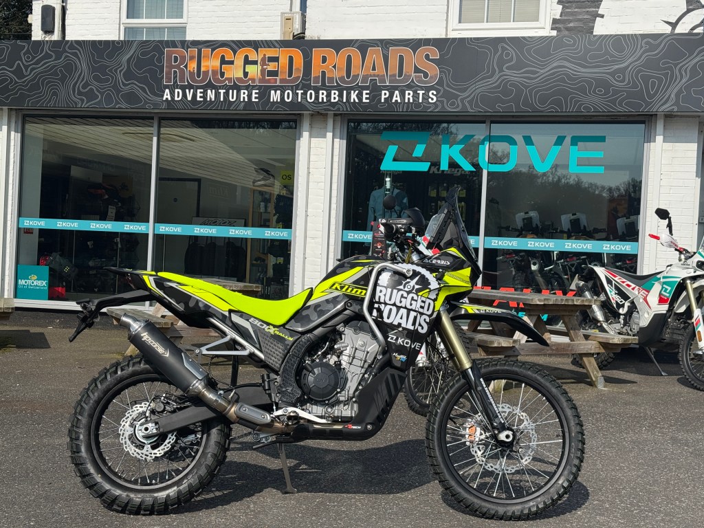 A bright yellow adventure motorcycle parked outside a shop called "Rugged Roads". The shop displays various motorbike parts and accessories, with a sign prominently featuring the store's name.