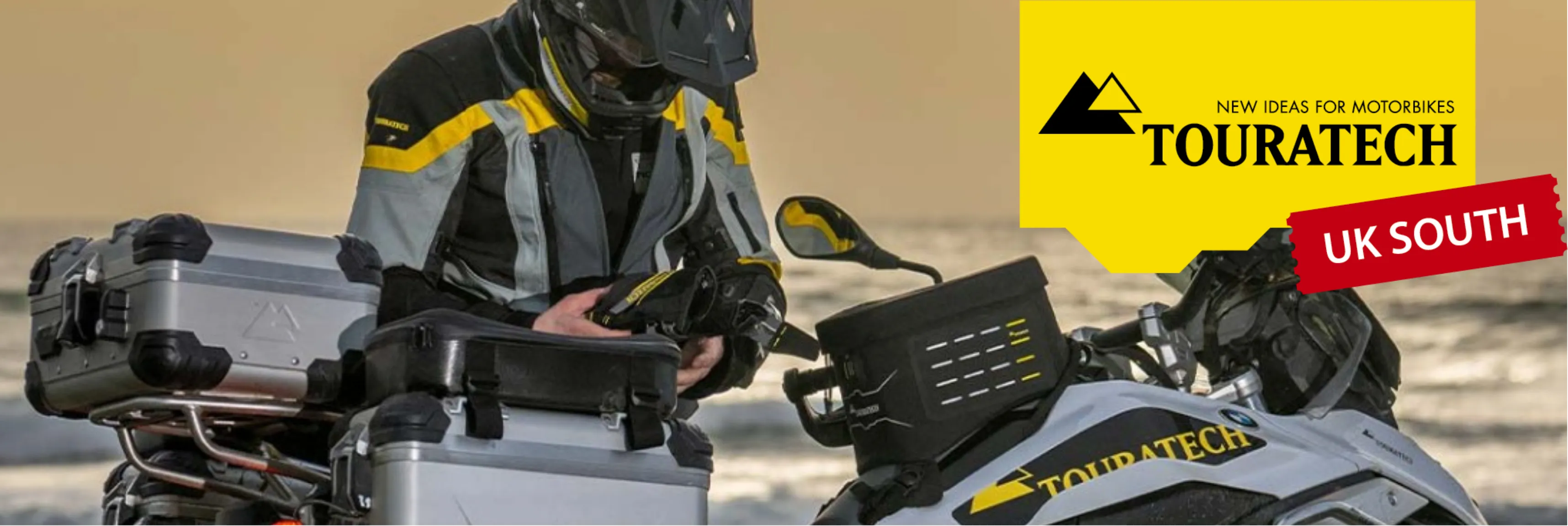 A motorcyclist in gear checking equipment on a BMW motorcycle, with Touratech branding and luggage, set against a beach backdrop.